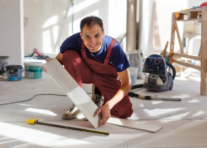 Ein Handwerker in Arbeitskleidung lächelt, während er Bodenfliesen oder -paneele in einem Raum verlegt, der renoviert wird.