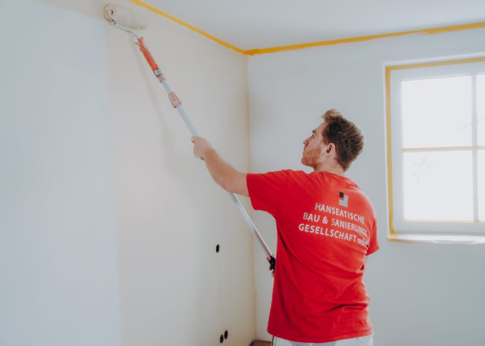 Ein Mitarbeiter der Hanseatischen Bau & Sanierungs Gesellschaft mbH streicht eine weiße Wand mit einer Farbrolle, Decke und Fenster sind abgeklebt. Maler Beruf