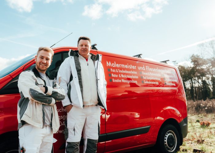 Zwei Handwerker in Arbeitskleidung stehen lächelnd vor einem roten Firmenwagen von HBS Lübeck, der Dienstleistungen als Malermeister und Fliesenlegerbetrieb bew
