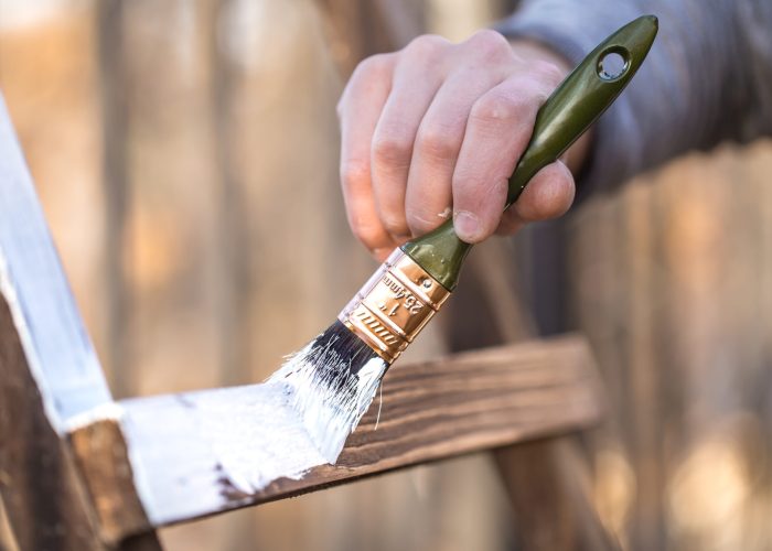 Eine männliche Hand streicht ein Holzobjekt mit weißer Farbe und einem Pinsel.