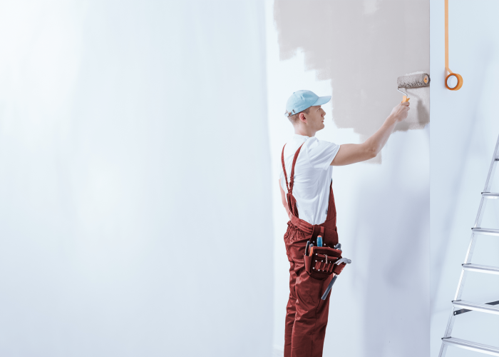 Ein Maler in roter Arbeitskleidung streicht eine Wand mit einer Farbrolle, während eine Leiter daneben steht.