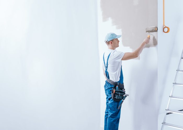 Ein Maler in blauer Arbeitskleidung streicht eine Wand mit einer Farbrolle, während er einen Werkzeuggürtel trägt.
